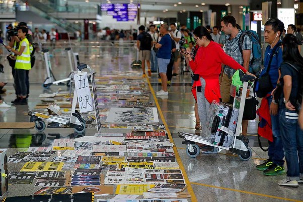 हांगकांग में हवाई अड्डे पर प्रदर्शनकारियों द्वारा लगाए गए तख्तों और पोस्टरों को देखते यात्री