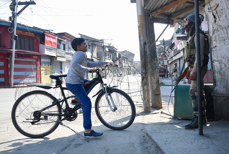 श्रीनगर में प्रतिबंधों के दौरान सुरक्षाकर्मी से बात करता साइकिल सवार एक किशोर